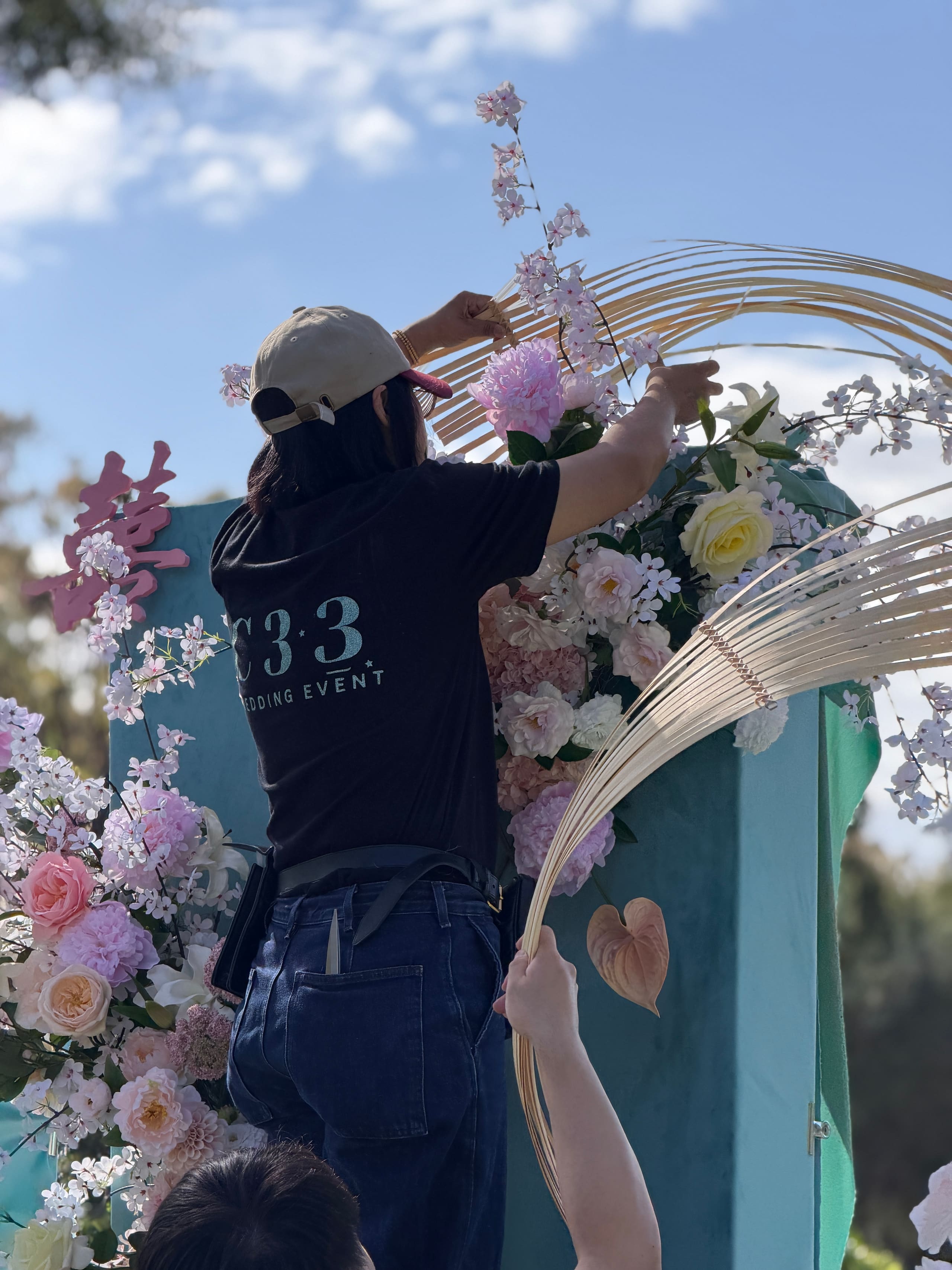 A C3 stylist installing pink florals and a rattan arch at an outdoor wedding setting