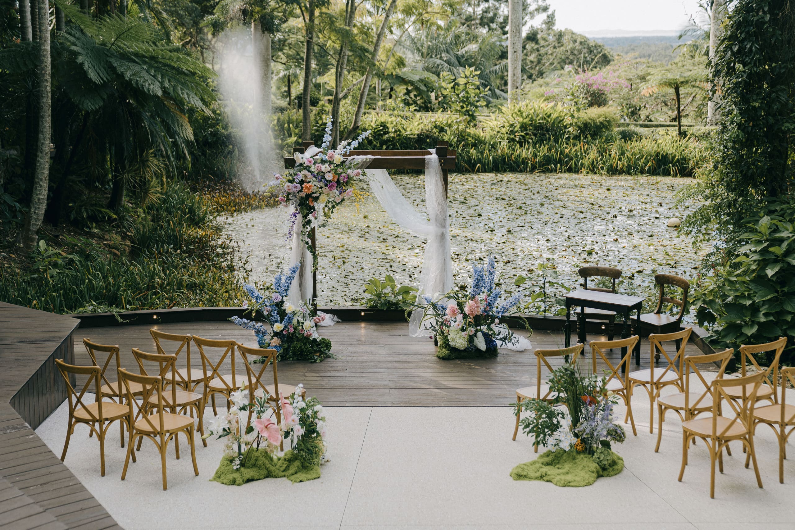 A garden ceremony beside a lily pond, with floral aisle arrangements and timber chairs