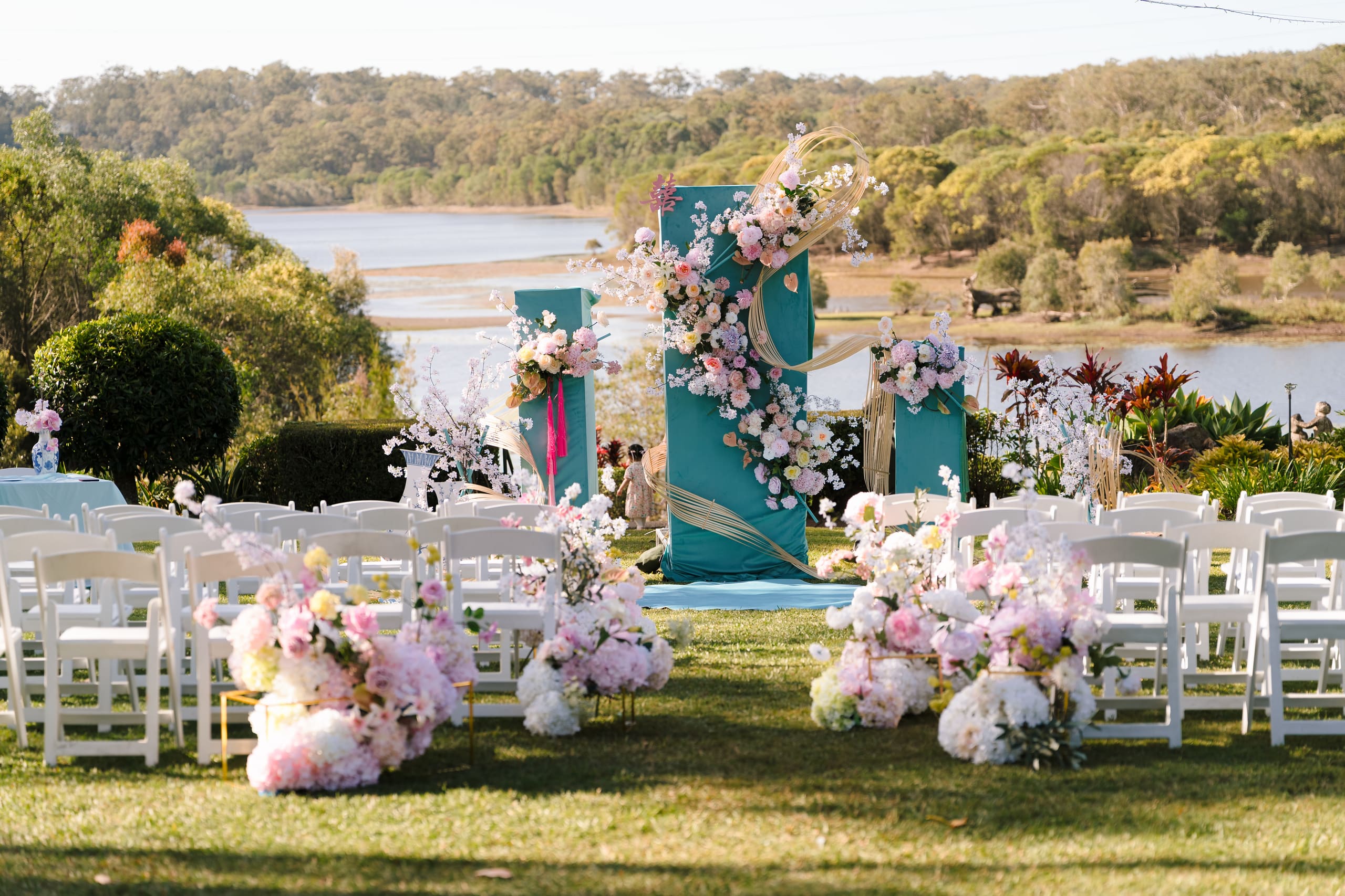 Lakeside ceremony setting with teal columns, pink and white floral installations and rows of white chairs