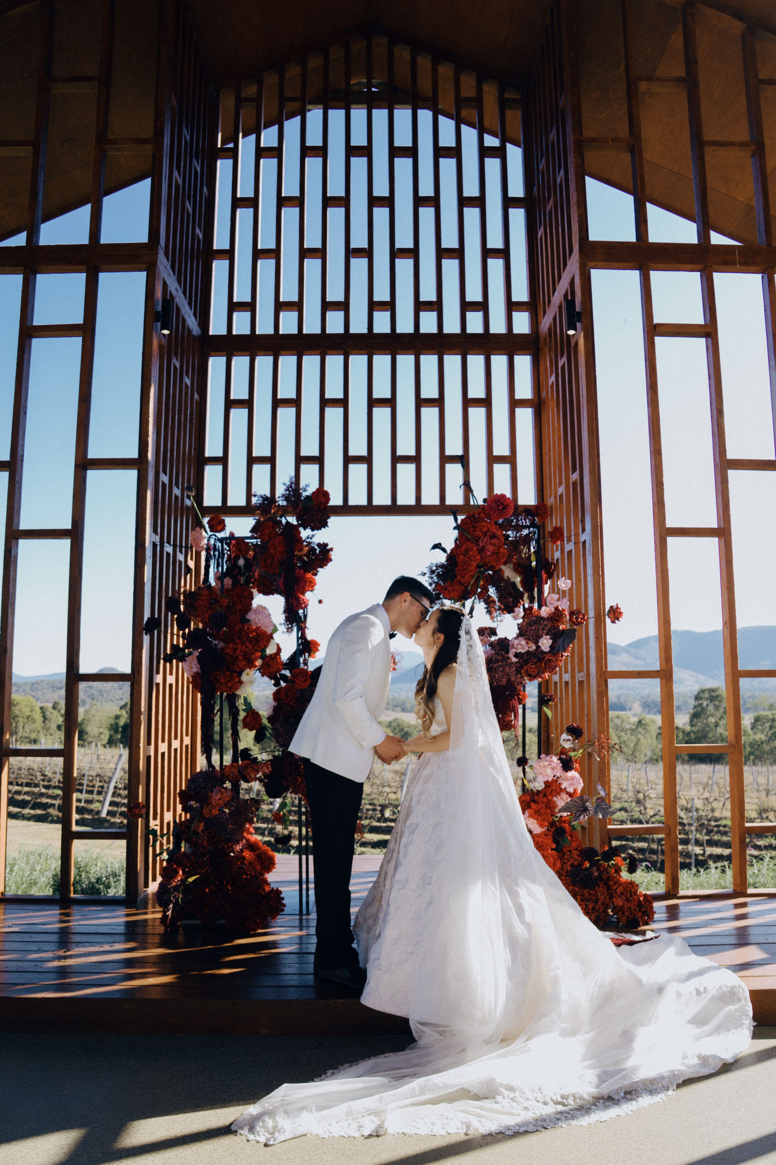 The couple beneath deep red floral installations in a timber and glass pavilion