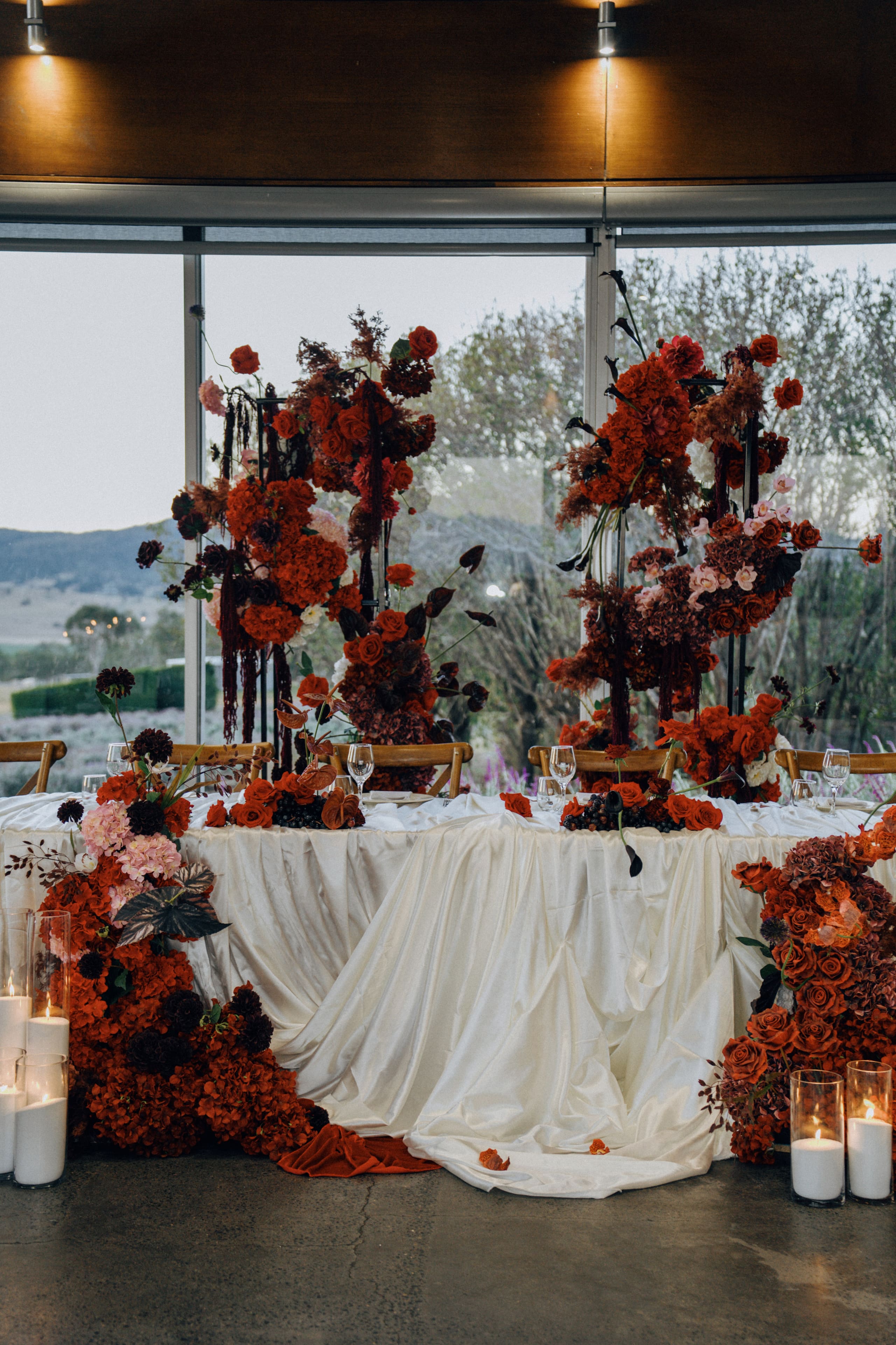 A head table framed by tall red and plum floral towers, draped ivory linen and candlelight